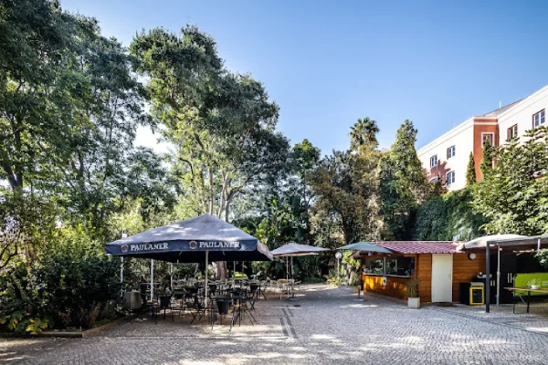 Il bar all'aperto con il Dehors del Goethe-Institut di Lisbona con tavoli e sedie ombreggiati sotto grandi ombrelloni. Il locale è circondato da alberi e verde, sulla destra è visibile un piccolo bancone in legno. Gli edifici sono parzialmente visibili sullo sfondo sotto un cielo azzurro.