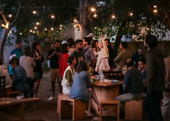 Un gruppo di persone in esposizioni si riunisce all'aperto, di notte, sotto le luci di Lisbona creando un'atmosfera vivace e calda tipica degli eventi o delle mostre. Alcuni sono in piedi e chiacchierano, altri sono seduti, socializzando attorno a tavoli di legno.