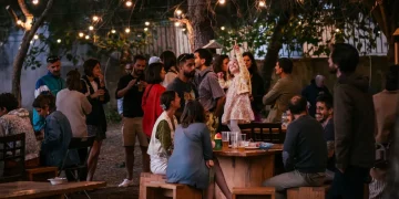 Un gruppo di persone in esposizioni si riunisce all'aperto, di notte, sotto le luci di Lisbona creando un'atmosfera vivace e calda tipica degli eventi o delle mostre. Alcuni sono in piedi e chiacchierano, altri sono seduti, socializzando attorno a tavoli di legno.