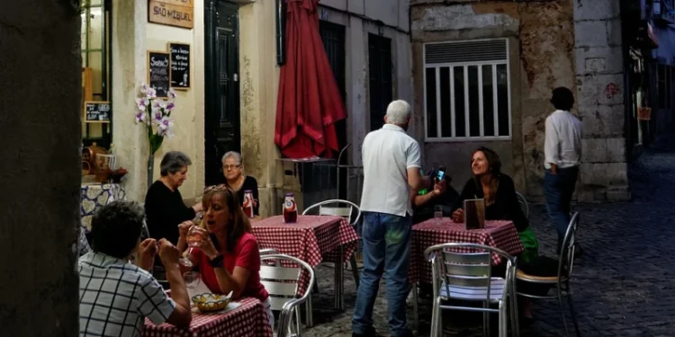 Le persone contribuiscono al fatturato sedendo ai tavoli all'aperto di un piccolo ristorante a Lisbona in Portogallo. Sono impegnati in conversazioni e pranzi sotto le decorazioni rosse appese sopra. Un cameriere sta in piedi mentre parla a un tavolo. Il ristorante ha un'atmosfera rustica con tovaglie a quadretti.