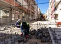 Operai arrivati con immigrazione che ripavimentano una strada acciottolata tra gli edifici in Portogallo. Un operaio è in ginocchio e sistema le pietre, mentre un altro manovra un veicolo da cantiere in lontananza. Attorno all'area di lavoro sono visibili barriere e strumenti di sicurezza. La luce del giorno brillante illumina la scena, mostrando il futuro dello sviluppo urbano.