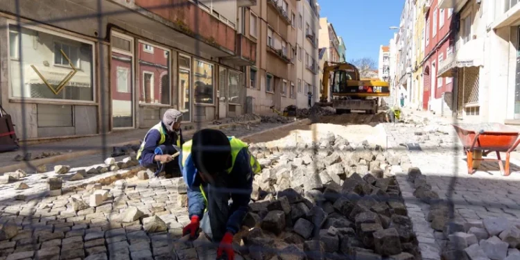 Operai arrivati con immigrazione che ripavimentano una strada acciottolata tra gli edifici in Portogallo. Un operaio è in ginocchio e sistema le pietre, mentre un altro manovra un veicolo da cantiere in lontananza. Attorno all'area di lavoro sono visibili barriere e strumenti di sicurezza. La luce del giorno brillante illumina la scena, mostrando il futuro dello sviluppo urbano.