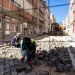 Operai arrivati con immigrazione che ripavimentano una strada acciottolata tra gli edifici in Portogallo. Un operaio è in ginocchio e sistema le pietre, mentre un altro manovra un veicolo da cantiere in lontananza. Attorno all'area di lavoro sono visibili barriere e strumenti di sicurezza. La luce del giorno brillante illumina la scena, mostrando il futuro dello sviluppo urbano.