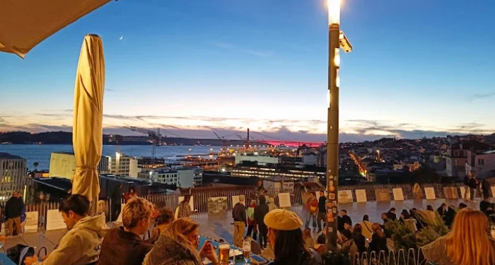 Un gruppo di persone siede su una delle migliori miradouri con bar all'aperto e dohors al tramonto, con vista su Lisbona e su il Tejo. Il cielo sta passando dal tramonto alla notte, con una falce di luna visibile. La scena comprende edifici urbani, gru e un ponte ben illuminato sullo sfondo.