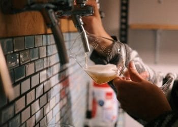 Siamo all'interno di una delle migliori birrerie artigianali di Lisbona una persona versa la birra dala spina in un bicchiere inclinato, formando una schiuma schiumosa nella parte superiore. L'accogliente area interna con pareti piastrellate e una mensola in legno emana un'atmosfera casual e rilassata.