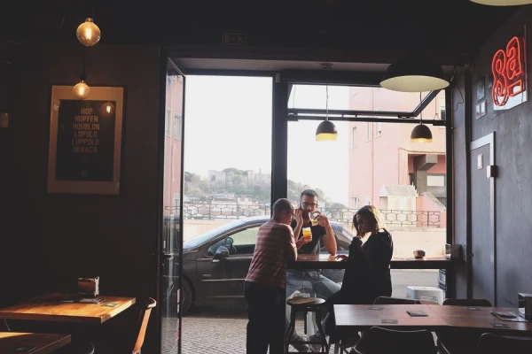 Scattata all'interno del pub 8a colina, una tra le migliori birrerie artigianali a Lisbona. Tre persone sono in piedi accanto a una porta aperta del bar poco illuminato, conversando e tenendo in mano una birra. All'esterno è parcheggiata un'auto con sullo sfondo i palazzi di Lisbona.