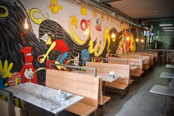 
L'interno di una delle migliori birrerie artigianali a Lisbona, nome Dois Corvos, è caratterizzato da lunghe panche in legno con ripiani in marmo. Le pareti sono adornate con graffiti colorati e giocosi che raffigurano vari personaggi dei cartoni animati e scene stravaganti. Le lampadine sospese in stile industriale si aggiungono all'estetica moderna.