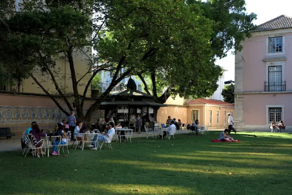 La gente è seduta ai tavolini all'aperto, all'ombra degli alberi, in un garden café, uno dei migliori dehors di Lisbona. Altri sono seduti sull'erba, godendosi una giornata di sole. Sullo sfondo sono visibili degli edifici, incluso uno con la facciata piastrellata. L'atmosfera appare rilassata e allegra.