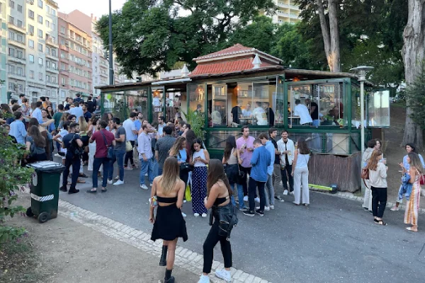 Una folla vivace si raduna all'esterno di un dei migliori dehors con grandi vetrate, il Corner Quiosque di Lisbona. Molti sono impegnati in una conversazione, mentre altri stanno in gruppo. Lo sfondo presenta edifici residenziali e alberi, che indicano un contesto urbano.