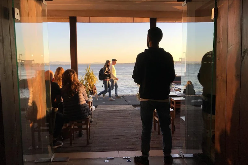 Una persona si trova in piedi osserva le migliori persone nel dehors del bar affacciato sul Tejo a Libsona. All'interno dell'edificio, diverse persone sono sedute a un tavolo vicino alla porta aperta, con la luce del sole che proietta toni caldi sulla scena. Lo sfondo è caratterizzato da uno specchio d'acqua calmo e da un affascinante allestimento del Dehors.