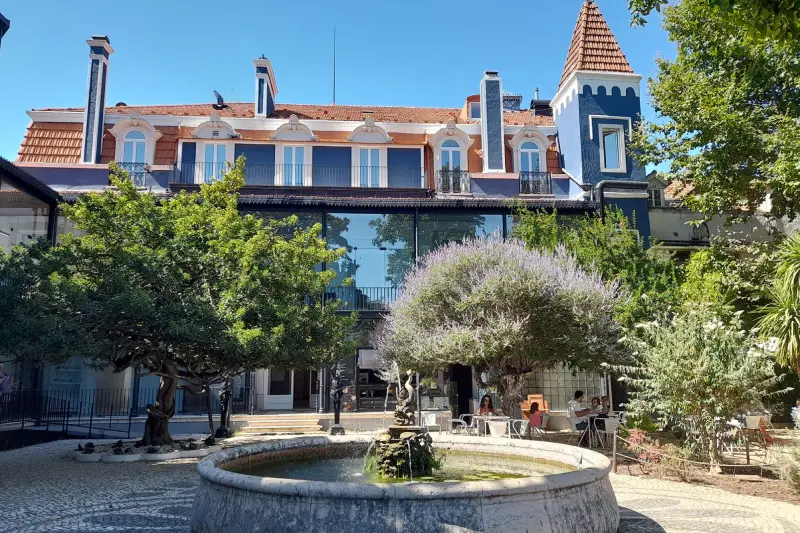 Uno dei Migliori di Dehors o Lisbona situato nell'incantevole cortile di Palácio Baldaya a Benfica. Si presenta con una fontana centrale in pietra circondata da una vegetazione lussureggiante, alberi e posti a sedere all'aperto. Sullo sfondo, sotto un cielo azzurro e limpido, si erge per tre piani un caratteristico edificio con il tetto in terracotta e una torretta blu.