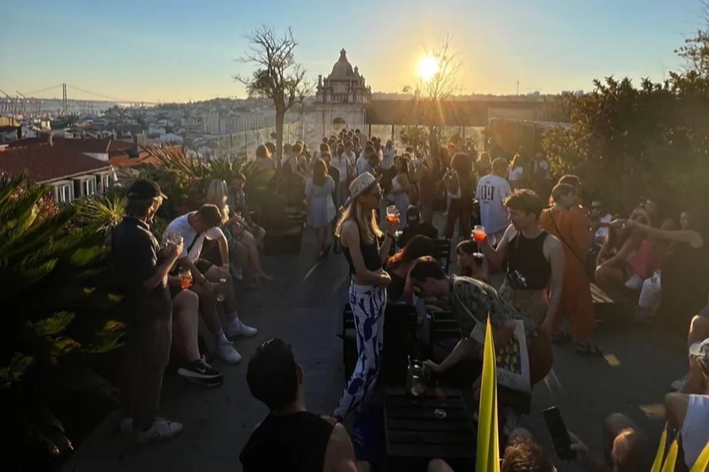 Uno dei migliori roof all'aperto quello del Park bar Lisbona, pieno di gente che si gode un drink e socializza durante il tramonto. La scena si affaccia su un paesaggio urbano con ponti ed edifici distanti. Le persone sono sedute e in piedi, indossano abiti estivi casual, mentre si immergono nell'atmosfera vibrante.