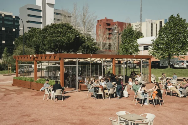 Uno de migliori Dehors quello del Quiosque Verde Lima di Lisbona, con persone sedute ai tavolini che gustano cibi e bevande sotto un cielo terso. La caffetteria dispone di un pergolato in legno ed è situata in una moderna area urbana con alti edifici sullo sfondo e alberi verdi nelle vicinanze.