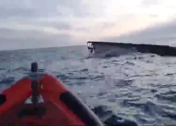 Dopo il naufragio a Marinha Grande il peschereccio Virgem Dolorosa parzialmente sommerso è visibile in lontananza con mare mosso e cielo nuvoloso. In primo piano si vede la prua di una barca di salvataggio gonfiabile arancione.