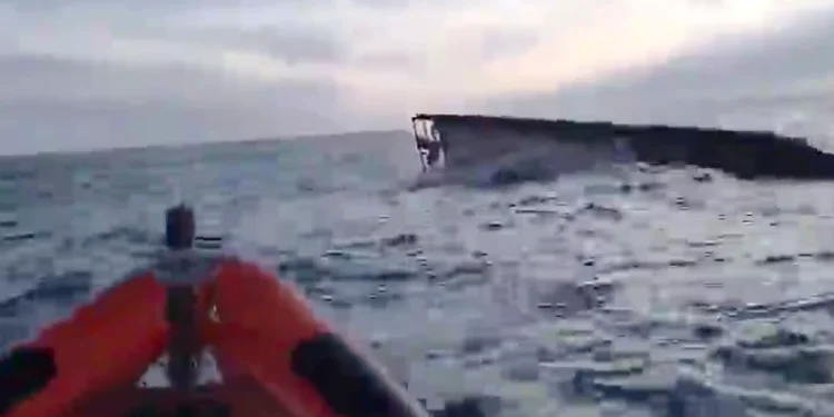 Dopo il naufragio a Marinha Grande il peschereccio Virgem Dolorosa parzialmente sommerso è visibile in lontananza con mare mosso e cielo nuvoloso. In primo piano si vede la prua di una barca di salvataggio gonfiabile arancione.