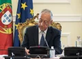Il presidente della repubblica portoghese è seduto a una scrivania decorata e firma il piano per una riduzione IRS. Dietro di lui ci sono le bandiere del Portogallo e dell'Unione Europea. Marcelo rebelo de Sousa appare concentrato sul suo lavoro, con microfoni e una bottiglia d'acqua sulla scrivania davanti a lui.