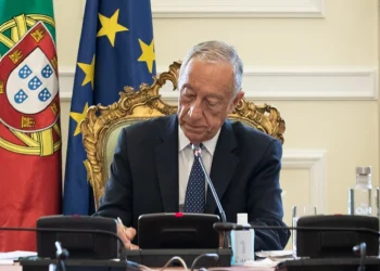 Il presidente della repubblica portoghese è seduto a una scrivania decorata e firma il piano per una riduzione IRS. Dietro di lui ci sono le bandiere del Portogallo e dell'Unione Europea. Marcelo rebelo de Sousa appare concentrato sul suo lavoro, con microfoni e una bottiglia d'acqua sulla scrivania davanti a lui.
