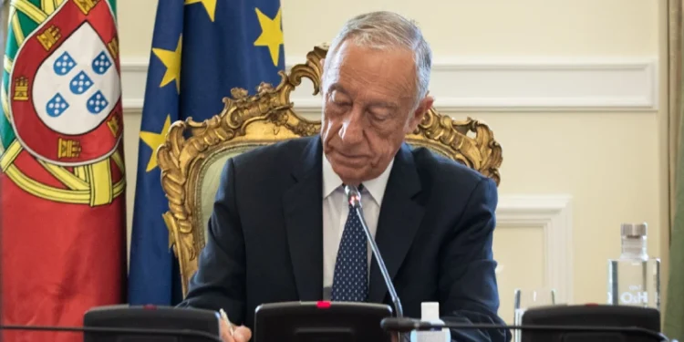 Il presidente della repubblica portoghese è seduto a una scrivania decorata e firma il piano per una riduzione IRS. Dietro di lui ci sono le bandiere del Portogallo e dell'Unione Europea. Marcelo rebelo de Sousa appare concentrato sul suo lavoro, con microfoni e una bottiglia d'acqua sulla scrivania davanti a lui.