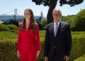La Principessa Leonor di Spagna e il Presidente della repubblica portoghese Marcelo rebelo de Sousa stanno fianco a fianco all'aperto durante la visita ufficiale a Lisbona. Stanno entrambi sorridendo. Sullo sfondo si trovano siepi verdi rigogliose e un albero, insieme a un grande ponte e altre strutture contro un cielo limpido.