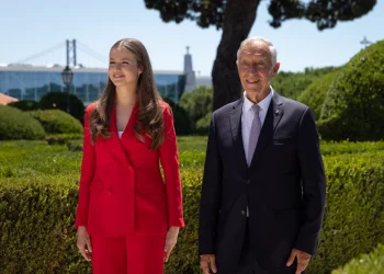 La Principessa Leonor di Spagna e il Presidente della repubblica portoghese Marcelo rebelo de Sousa stanno fianco a fianco all'aperto durante la visita ufficiale a Lisbona. Stanno entrambi sorridendo. Sullo sfondo si trovano siepi verdi rigogliose e un albero, insieme a un grande ponte e altre strutture contro un cielo limpido.