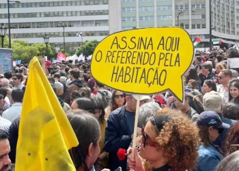 Una grande folla di persone partecipano ad una manifestazione per richiedere un referendum contro gli AL. Un manifestante tiene in mano un cartello giallo brillante con la scritta "Assina Aqui O Referendo Pela Habitação", che si traduce in "Firma qui per il referendum sulla casa". Il raduno mette in luce temi come gli affitti delle case ai turisti e le raccolte le firme.