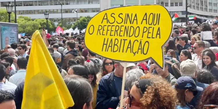 Una grande folla di persone partecipano ad una manifestazione per richiedere un referendum contro gli AL. Un manifestante tiene in mano un cartello giallo brillante con la scritta "Assina Aqui O Referendo Pela Habitação", che si traduce in "Firma qui per il referendum sulla casa". Il raduno mette in luce temi come gli affitti delle case ai turisti e le raccolte le firme.