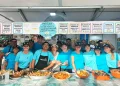 Un gruppo di persone dello staff della "Festa da Ria Formosa" sta dietro un bancone delle sagre del pesce in Algarve, Portogallo. Stanno sorridendo e posando con vari piatti di pesce sul bancone di fronte a loro, tra cui paella, crostacei e aragosta. I menu del cibo sono visualizzati sopra di loro.