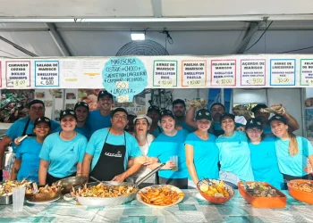Un gruppo di persone dello staff della "Festa da Ria Formosa" sta dietro un bancone delle sagre del pesce in Algarve, Portogallo. Stanno sorridendo e posando con vari piatti di pesce sul bancone di fronte a loro, tra cui paella, crostacei e aragosta. I menu del cibo sono visualizzati sopra di loro.