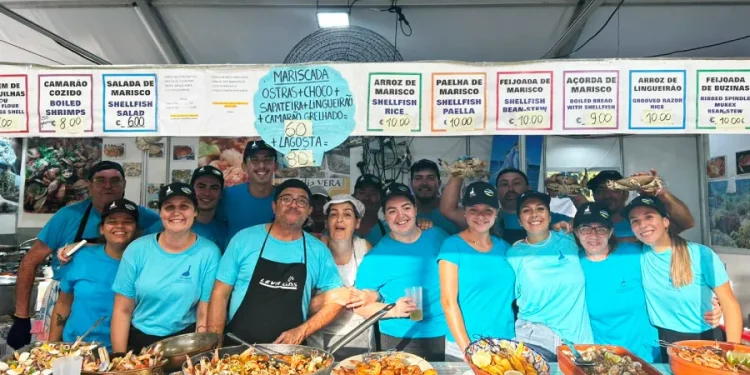 Un gruppo di persone dello staff della "Festa da Ria Formosa" sta dietro un bancone delle sagre del pesce in Algarve, Portogallo. Stanno sorridendo e posando con vari piatti di pesce sul bancone di fronte a loro, tra cui paella, crostacei e aragosta. I menu del cibo sono visualizzati sopra di loro.