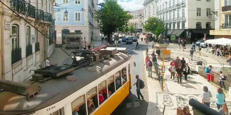 Scena di strada in una vivace Lisbona, con un tram giallo, con turisti che pagano la tassa di soggiorno a Lisbona. La gente cammina sui marciapiedi e attraversa la strada con caffè all'aperto ed edifici storici che fiancheggiano la strada sotto un cielo azzurro, parzialmente nuvoloso.