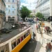Scena di strada in una vivace Lisbona, con un tram giallo, con turisti che pagano la tassa di soggiorno a Lisbona. La gente cammina sui marciapiedi e attraversa la strada con caffè all'aperto ed edifici storici che fiancheggiano la strada sotto un cielo azzurro, parzialmente nuvoloso.