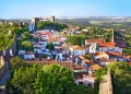 Una vista panoramica della cose da vedere nella città storica a Obidos con edifici bianchi e tetti di tegole rosse racchiusi da antiche mura in pietra. La città è circondata da rigogliosi alberi verdi e dolci colline sotto un cielo azzurro e terso.