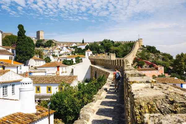 Una delle cose da vedere nella città storica a Obidos con la vista panoramica sulla città medievale fortificata con mura di pietra che si estendono in lontananza sormontate da un castello. Le persone camminano lungo lo stretto sentiero sopraelevato accanto a case rustiche con tetti di tegole rosse, incorniciate da una vegetazione lussureggiante sotto un cielo parzialmente nuvoloso.