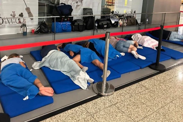 Turisti Italiani sono sdraiati su materassini blu in un'area interna separata da una barriera di corda rossa. Avvolti in coperte blu, sembrano riposare o dormire, con borse e oggetti personali riposti contro un muro dietro di loro. Il pavimento è piastrellato con un motivo a macchie.