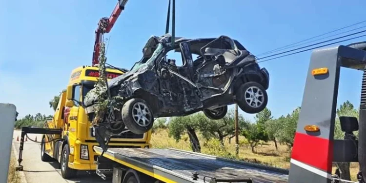 Un'auto gravemente danneggiata viene sollevata da una gru sul retro di un carro attrezzi dopo un incidente stradale su una strada rurale in Portogallo. L'auto è gravemente schiacciata. Un altro carro attrezzi è parcheggiato lì vicino. La scena è ambientata sotto un cielo azzurro e terso con alberi sullo sfondo.