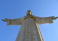 Una statua del Cristo Rei a Lisbona, con le braccia tese, con Porto sono le città più invase dai turisti in Europa. Vista dal basso, la grandiosità della statua e la lavorazione dettagliata della sua veste e dei lineamenti del viso affascinano sia la gente del posto che i turisti. Sullo sfondo un cielo sereno di colore blu.