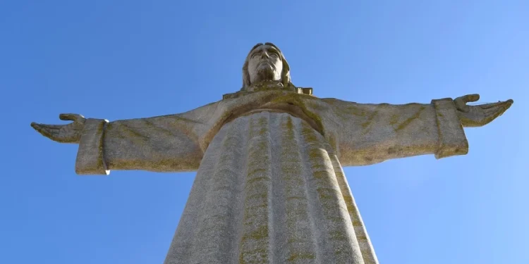 Una statua del Cristo Rei a Lisbona, con le braccia tese, con Porto sono le città più invase dai turisti in Europa. Vista dal basso, la grandiosità della statua e la lavorazione dettagliata della sua veste e dei lineamenti del viso affascinano sia la gente del posto che i turisti. Sullo sfondo un cielo sereno di colore blu.
