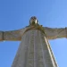 Una statua del Cristo Rei a Lisbona, con le braccia tese, con Porto sono le città più invase dai turisti in Europa. Vista dal basso, la grandiosità della statua e la lavorazione dettagliata della sua veste e dei lineamenti del viso affascinano sia la gente del posto che i turisti. Sullo sfondo un cielo sereno di colore blu.