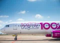 Un aereo Wizz Air è parcheggiato su una pista di atterraggio soleggiata, evidenziando il loro All You Can Fly abbonamento volo per soli 499€ all'anno. Si legge la scritta "Wizz 100" in grandi lettere rosa e viola sulla fusoliera. Piccole nuvole bianche punteggiano il cielo azzurro brillante sullo sfondo.