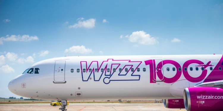 Un aereo Wizz Air è parcheggiato su una pista di atterraggio soleggiata, evidenziando il loro All You Can Fly abbonamento volo per soli 499€ all'anno. Si legge la scritta "Wizz 100" in grandi lettere rosa e viola sulla fusoliera. Piccole nuvole bianche punteggiano il cielo azzurro brillante sullo sfondo.