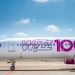 Un aereo Wizz Air è parcheggiato su una pista di atterraggio soleggiata, evidenziando il loro All You Can Fly abbonamento volo per soli 499€ all'anno. Si legge la scritta "Wizz 100" in grandi lettere rosa e viola sulla fusoliera. Piccole nuvole bianche punteggiano il cielo azzurro brillante sullo sfondo.