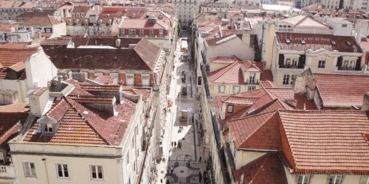 Una vista aerea di Lisbona con strade strette e palazzi fitti e dai tetti rossi, stando all'andamento degli affitti in Portogallo. Le strade sono piene di pedoni e fiancheggiate da edifici a più piani, dando un senso di densità urbana e uniformità architettonica.