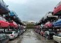 Un deposito con numerose file di auto accatastate e abbandonate di vari colori e condizioni. I veicoli, molti dei quali hanno superato di gran lunga l'età media delle auto in Portogallo, sono disposti in file, con una fila sopra l'altra, che si estendono in lontananza. Il terreno è bagnato, riflettendo il cielo coperto sopra.