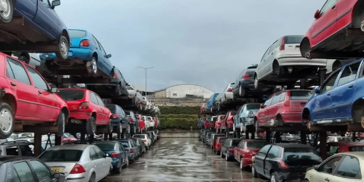 Un deposito con numerose file di auto accatastate e abbandonate di vari colori e condizioni. I veicoli, molti dei quali hanno superato di gran lunga l'età media delle auto in Portogallo, sono disposti in file, con una fila sopra l'altra, che si estendono in lontananza. Il terreno è bagnato, riflettendo il cielo coperto sopra.