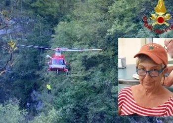 Un elicottero di soccorso rosso volteggia vicino a una rupe boscosa, mentre recupera una persona, nel riquadro è inserita la foto di Fiorenza Chiesa morta per la caduta nel bosco. L'emblema dei vigili del fuoco italiani è nell'angolo in alto a destra.