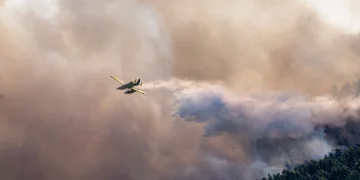 Un aereo antincendio rilascia acqua su un incendio boschivo che si sta diffondendo in una fitta foresta nel nord del Portogallo. Un fumo denso riempie il cielo e una parte della volta degli alberi è visibile sotto le nuvole di fumo che si sollevano dall'incendio.