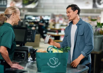 Un cliente alla cassa di un supermercato mostra il suo cellulare a una cassiera, entrambi sorridenti e con in mano una borsa della spesa riutilizzabile piena di generi alimentari, è entusiasta di usare un'app incentrata su Spreco Alimentare. La app è Too Good To Go.