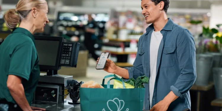 Un cliente alla cassa di un supermercato mostra il suo cellulare a una cassiera, entrambi sorridenti e con in mano una borsa della spesa riutilizzabile piena di generi alimentari, è entusiasta di usare un'app incentrata su Spreco Alimentare. La app è Too Good To Go.