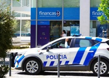 Un'auto della polizia blu e bianca è parcheggiata di fronte a un edificio per indagare sulle truffe alla Finanças tramite email. Il palazzo ha una facciata in vetro e la scritta "Finanças". L'edificio, contrassegnato da una segnaletica blu, funge da ufficio dell'autorità fiscale e doganale. Vicino all'ingresso c'è un individuo che passa. Alberi e dissuasori sono in primo piano.