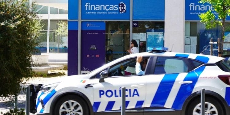 Un'auto della polizia blu e bianca è parcheggiata di fronte a un edificio per indagare sulle truffe alla Finanças tramite email. Il palazzo ha una facciata in vetro e la scritta "Finanças". L'edificio, contrassegnato da una segnaletica blu, funge da ufficio dell'autorità fiscale e doganale. Vicino all'ingresso c'è un individuo che passa. Alberi e dissuasori sono in primo piano.
